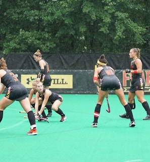 Several athletes in matching sports uniforms are on a bright green field, engaged in a field hockey game. They appear focused and are holding hockey sticks, with some bending slightly as they prepare to play. Trees and a billboard are visible in the background.