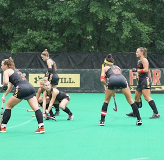 Several athletes in matching sports uniforms are on a bright green field, engaged in a field hockey game. They appear focused and are holding hockey sticks, with some bending slightly as they prepare to play. Trees and a billboard are visible in the background.