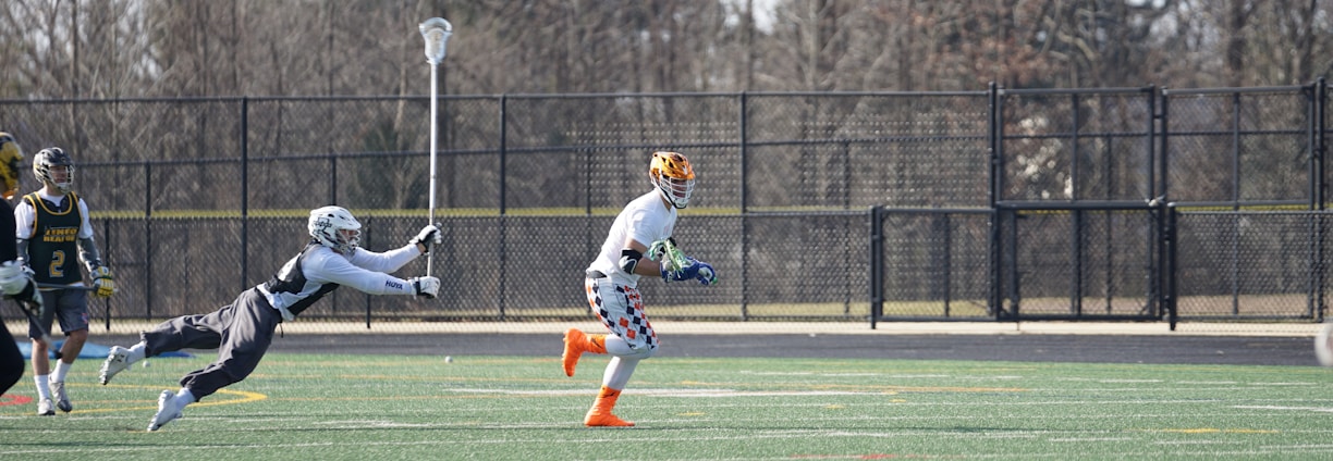 A group of athletes engaged in a sports game on a green field. One player is running forward with possession of the ball, wearing colorful socks and a checkered uniform, while another player in dark clothing dives to intercept or tackle. Other players are present in the background. Leafless trees and a metal fence surround the field.