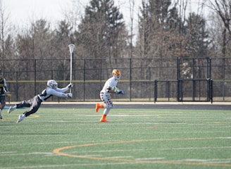A group of athletes engaged in a sports game on a green field. One player is running forward with possession of the ball, wearing colorful socks and a checkered uniform, while another player in dark clothing dives to intercept or tackle. Other players are present in the background. Leafless trees and a metal fence surround the field.