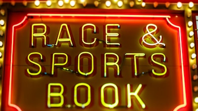Bright neon lights on a marquee sign display the words 'RACE & SPORTS BOOK'. The sign is surrounded by rows of yellow and white lights against a red background, evoking a vibrant, energetic atmosphere typically associated with casinos or entertainment venues.