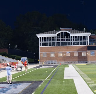 A nighttime scene featuring a sports stadium with a partially filled seating area and a large moon visible in the sky. The field is mostly empty except for a few people scattered around, and there are bright lights illuminating the venue. The stadium is surrounded by trees and a prominent sign displaying 'Maryland Lottery' is visible.