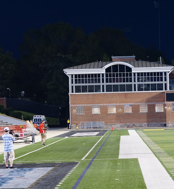 A nighttime scene featuring a sports stadium with a partially filled seating area and a large moon visible in the sky. The field is mostly empty except for a few people scattered around, and there are bright lights illuminating the venue. The stadium is surrounded by trees and a prominent sign displaying 'Maryland Lottery' is visible.