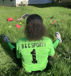 A person with curly hair sits on green grass facing away, wearing a bright green sports jersey with 'B & G SPORTS' and the number 1 on the back. Sports equipment, including a red helmet and baseballs, is scattered around.