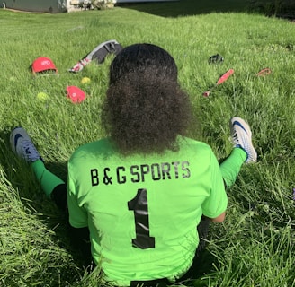 A person with curly hair sits on green grass facing away, wearing a bright green sports jersey with 'B & G SPORTS' and the number 1 on the back. Sports equipment, including a red helmet and baseballs, is scattered around.