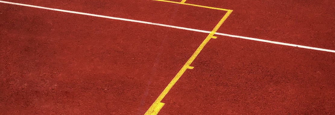A sports court with a reddish-brown surface and bright yellow and white lines. The lines form geometric shapes, including rectangles and circles, suggesting markings for games like basketball or netball.