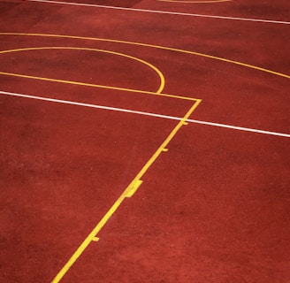 A sports court with a reddish-brown surface and bright yellow and white lines. The lines form geometric shapes, including rectangles and circles, suggesting markings for games like basketball or netball.