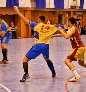 A dynamic indoor sports scene with players in motion on a court. Two athletes in blue and yellow uniforms are engaged in an intense handball game, with one player gripping the other's jersey. The background shows other players, several seated people, and advertisements on the walls.