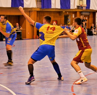 A dynamic indoor sports scene with players in motion on a court. Two athletes in blue and yellow uniforms are engaged in an intense handball game, with one player gripping the other's jersey. The background shows other players, several seated people, and advertisements on the walls.