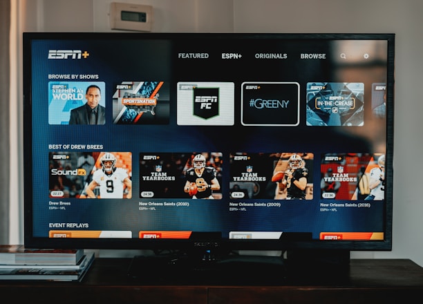 A television screen displaying the ESPN+ streaming interface with various sports-related shows and categories. The screen includes sections such as 'Browse by Shows' and 'Best of Drew Brees' with thumbnails of sports programs featuring athletes in action and analysts.