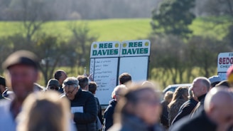 A group of people gathered outdoors with two large betting boards in the background labeled 'REG DAVIES'. The setting appears to be a countryside location with trees and fields visible in the distance. The crowd consists of individuals dressed in jackets and hats, engaging in conversation.