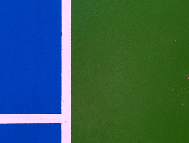 A close-up view of a sports court with a bright blue section on the left and a green section on the right, separated by white lines.