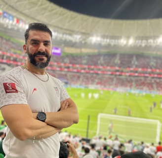 A person stands in a stadium filled with spectators during a sporting event. The grass field and goalposts are visible in the background. The individual is wearing a white jersey with a sports logo and is smiling at the camera.