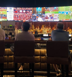 A group of people sit at a bar with their backs to the camera, watching multiple sports games displayed on large screens. The bar counter is lined with various bottles of liquor, and the atmosphere suggests a casual and relaxed social setting.