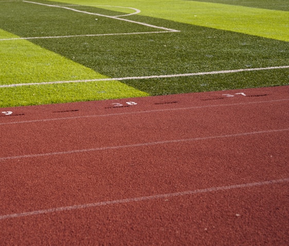 A sports field with vivid green grass divided into sections by white lines in the background. In the foreground, a track with a reddish-brown surface is marked with numbers and lines.