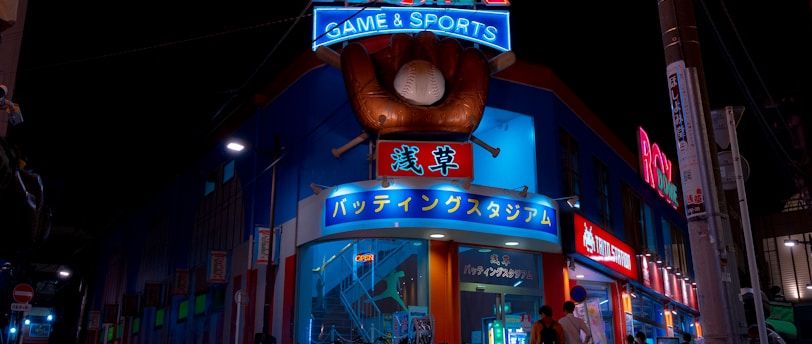 A vibrant, neon-lit facade of a gaming and sports establishment at night, featuring a large glove holding a baseball above the entrance. The building is adorned with colorful signs in both English and Japanese, with various advertisements and an illuminated sign that says 'ROX DOME'. Several people are walking towards the entrance, and bicycles are parked along the side.