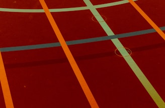 A sports court with intersecting lines in orange and blue, creating geometric patterns on a reddish-brown surface.