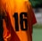 A person is wearing an orange sports jersey with the number 16 printed in black on the back. The background is blurred, but it appears to be an outdoor environment, possibly a sports field.