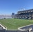 A large, empty sports stadium with blue seats surrounding a well-maintained green grass field. Bright sunlight creates clear shadows, and there are two goal posts on opposite ends of the field. A big electronic scoreboard displays some graphics, and the sky is clear and blue.