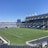 A large, empty sports stadium with blue seats surrounding a well-maintained green grass field. Bright sunlight creates clear shadows, and there are two goal posts on opposite ends of the field. A big electronic scoreboard displays some graphics, and the sky is clear and blue.